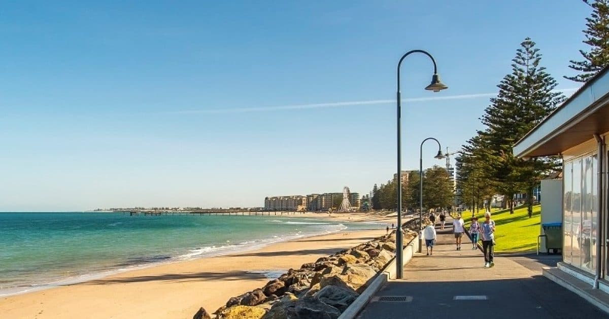 Beach esplanade in Adelaide with lampposts and pines, people walking beside the sand.