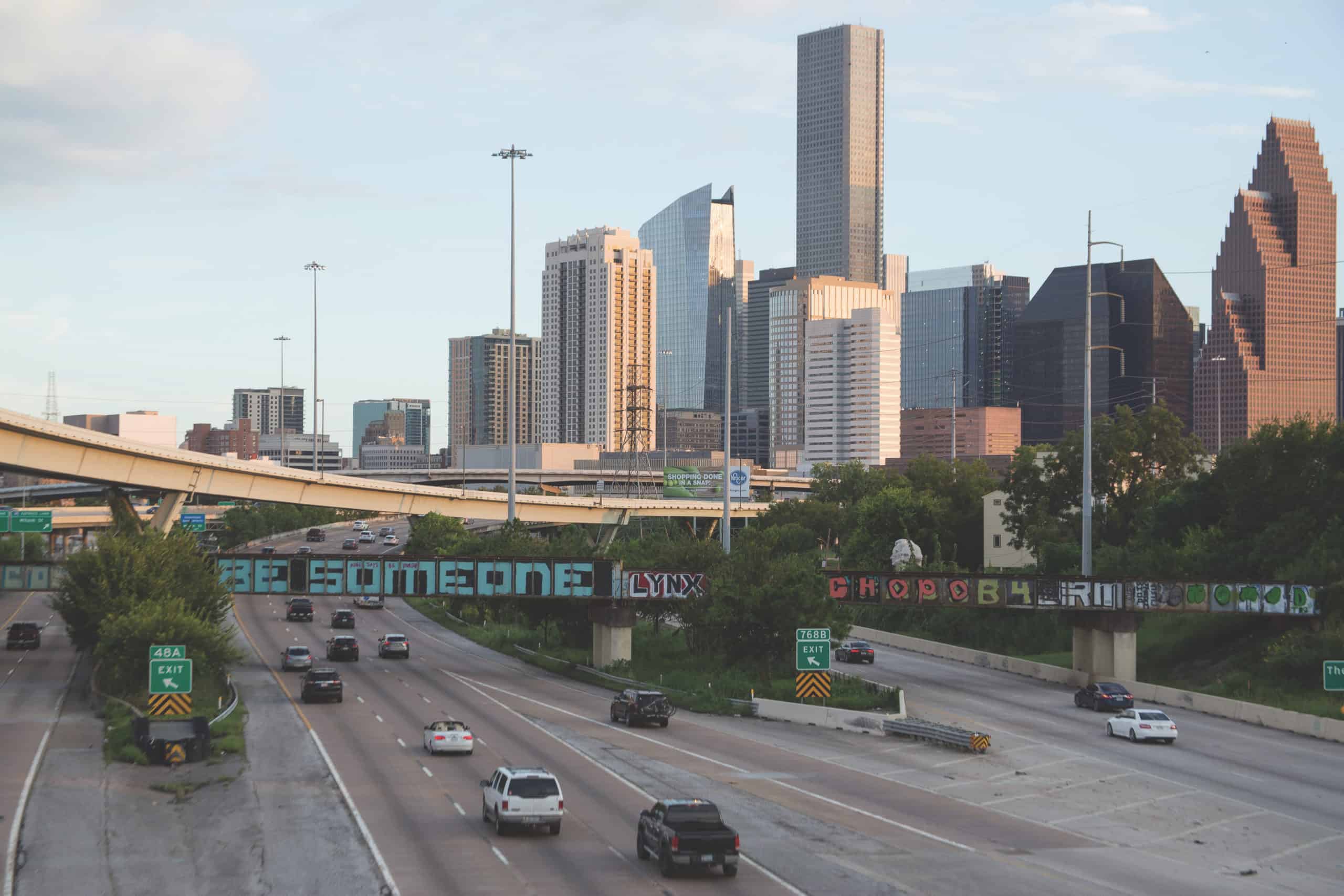 Houston skyline behind a rail bridge painted with the words BE SOMEONE above the freeway.